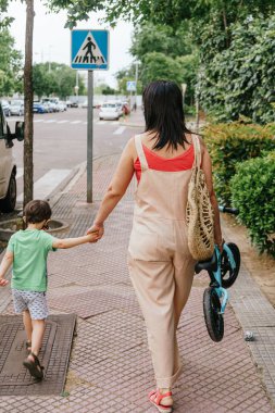 Anne ve oğlu kaldırımda beraber yürürler. O küçük bir bisiklet taşıyor. - Aile. Normallik