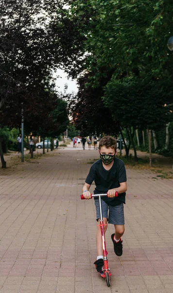 Askeri renkte sağlık maskesi takan, yaya yolunda scooter süren ve şort giyen çok ciddi bir çocuk. yeni normallik. Çocukluk. Paten süren bir çocuk. Çocuk dışarıda motosikletle oynuyor. Çocuk için aktif eğlence ve açık hava sporu