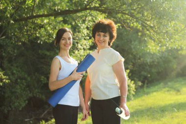 Olgun anne-kız spor paspası tutarak dışarıda spor yapmaya gidecekler. Aktif yaşam tarzı konsepti. Salgından sonra normal hayata yeniden başlamak.