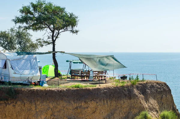 A house on wheels and a canopy under a tree on a cliff by the sea.
