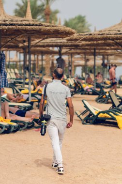 Deniz sahili boyunca yürüyen kameralı bir adam.