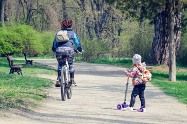 Scooter'lı küçük bir kız bisikletle yoldan geçen bir kıza bakıyor.
