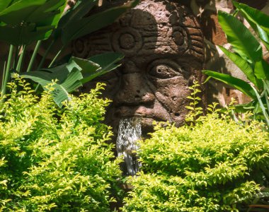 İlkbaharda bir halk parkında, taştan bir yüzün ağzından bir şelale çeşmesi geliyor..