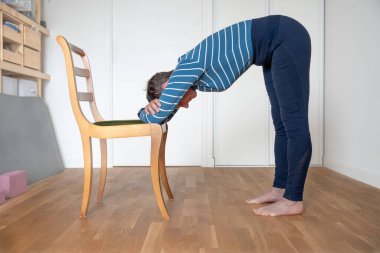A mature woman wearing a blue and white striped t-shirt and a blue gym shorts does yoga session at home