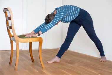 A mature woman wearing a blue and white striped t-shirt and a blue gym shorts does yoga session at home