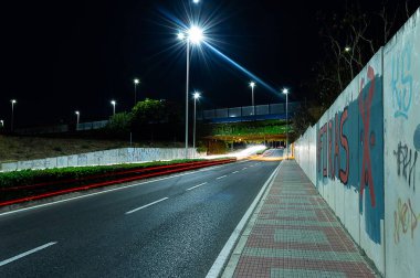 Boş yol yüzeyi, geceleri araba ışıkları ve ağaç ve bitki örtüsü olan bir köprü, duvar ve grafiti olan bir duvar.