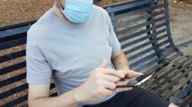 Beyaz bir genç adam parkta bankta dinleniyor ve mavi koruyucu bir maske takmış bir şekilde parmağıyla telefonu hareket ettiriyor. Sonbahar mevsimi