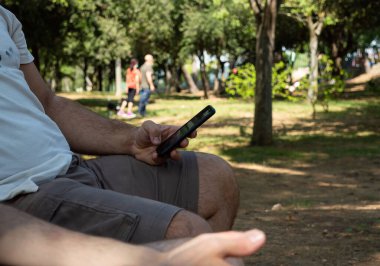 Yazın güneşli bir gününde doğa parkında akıllı telefon kullanan bir adam arka planda bulanıklaşır.