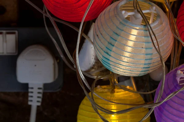 Colorful paper lantern fairy lights close up plugged into wall. Switched on colourful light & dark.