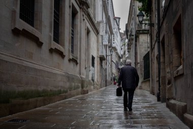 Santiago de Compostela, Galiçya, İspanya - 09 / 26 / 2020: Gri yağmurlu bir günde eski Santiago de Compostela kasabasında yalnız yaşlı bir adam bir çanta taşıyor. Yürüyen yaşlı adam, yalnız başına..