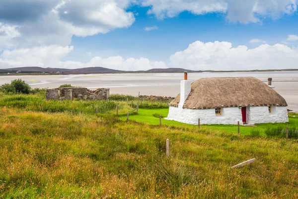 Chalet blanc traditionnellement construit avec toit de chaume, à côté de la baie turquoise, avec un ciel sombre nuageux orageux au-dessus. Île de North Uist, Écosse — Photo