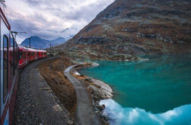 Bernina Express panoramic train passing by turquoise alpine lake in the Swiss Alps, scenic railway journey through mountains and valleys.