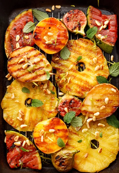 Grilled fruits; pineapples, peaches, figs, pears and watermelon with pine nuts, fresh herbs and honey on the grill plate, top view.