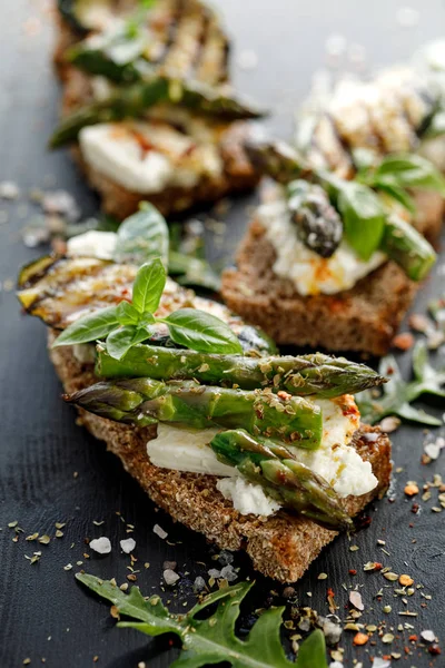 Vegetarian sandwich. Wholemeal bread sandwiches with feta cheese, grilled zucchini, green asparagus, sugar peas, olive oil and herbs on a dark background, close-up. Healthy eating concept