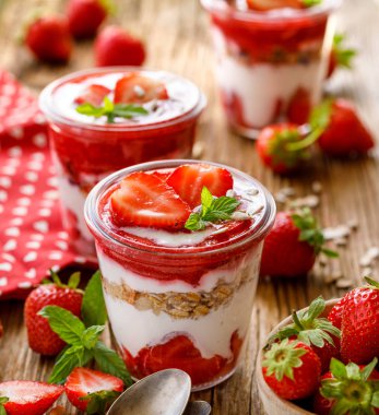 Çilek katmanlı tatlı, taze çilekli sağlıklı tatlı, doğal yoğurt, çilekli mus ve ahşap masadaki cam tabaklarda granola. Lezzetli ve besleyici tatlı ya da kahvaltı