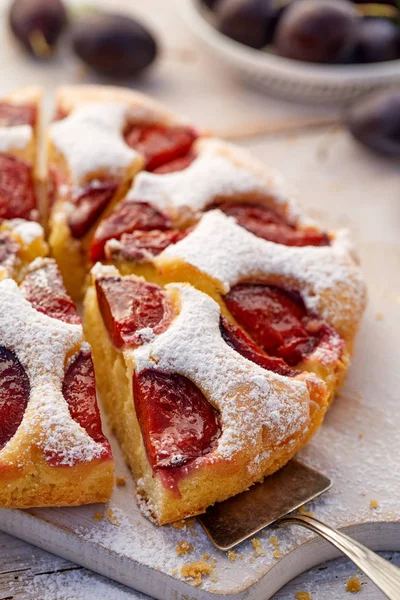 Plum cake, traditional homemade  cake with fruit divided into parts, sprinkled with powdered sugar on a white board, close-up view. Fruit cake, dessert