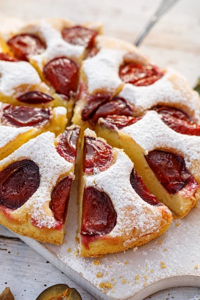 Plum cake, traditional homemade  cake with fruit divided into parts, sprinkled with powdered sugar on a white board, close-up view. Fruit cake, dessert