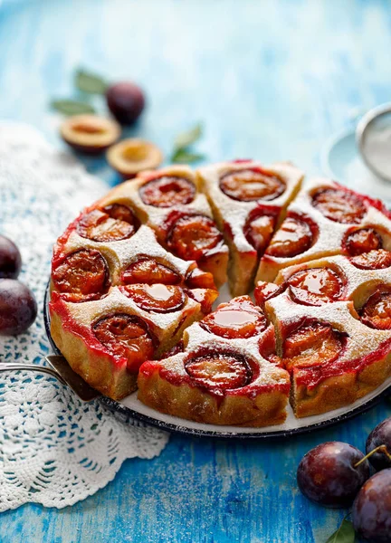 Plum cake, traditional homemade  cake with fruit, divided into portions, sprinkled with powdered sugar on a blue table, close-up view. Delicious fruitcake
