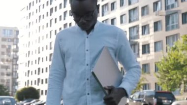Kendine güvenen genç siyahi işadamının portresi elinde dizüstü bilgisayarla kameraya bakıyor ve sokaklara gözlük takıyor, çalışmaya hazır, iş kavramı.