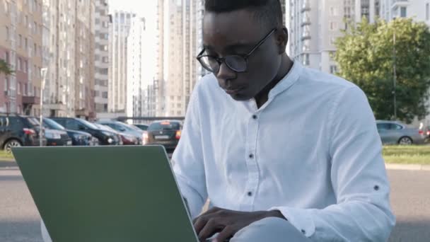 Empresario africano enfocado en escribir en la computadora portátil haciendo investigación. Joven profesional masculino usando computadora al aire libre. Trabajador freelancer ocupado trabajando distante en el dispositivo portátil de tecnología moderna — Vídeos de Stock