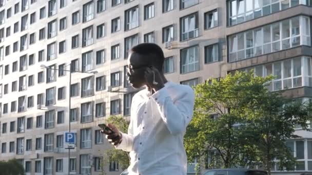 Hombre de negocios afroamericano joven guapo con auriculares haciendo danza de la victoria sobre el logro exitoso, hombre negro escuchando música y bailando activamente solo al aire libre cerca del edificio de oficinas — Vídeos de Stock