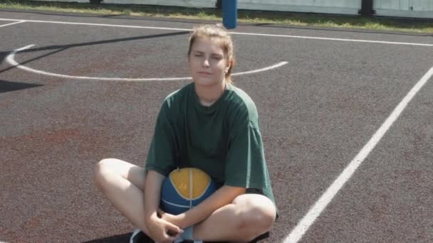 Zmęczony szczęśliwy koszykarka kobieta siedzi z piłką na boisku na świeżym powietrzu, odpoczywając — Wideo stockowe