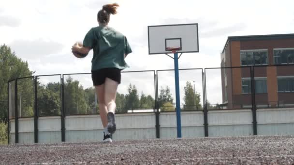 Баскетболистка в шортах и белых носках на профессиональной площадке бегает с мячом и успешно отбивает мяч к сетке. Запись с ручной клади. На открытом воздухе — стоковое видео