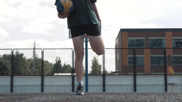 Спортивная стройная молодая баскетболистка, тренирующаяся на площадке под открытым небом, тренирующаяся перед баскетбольным матчем, занимающаяся спортом, сгибающая мышцы ног — стоковое видео