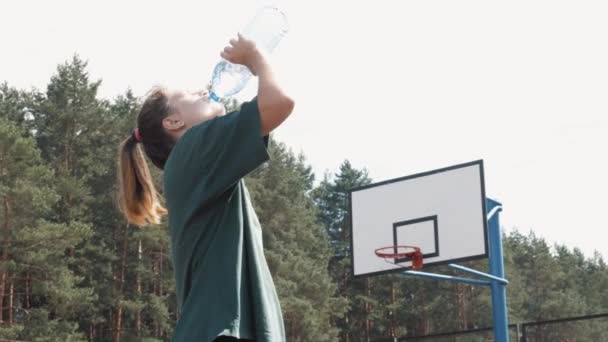 Потная уставшая баскетболистка пьет свежую холодную воду из пластиковой бутылки, измученная девочка-подросток во время спортивных соревнований — стоковое видео