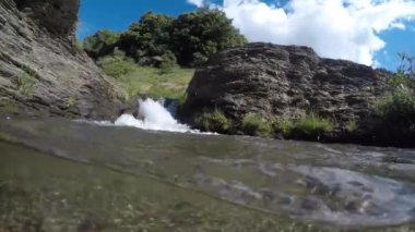 Etrafında bitki örtüsü olan su akıntısı. Kayalar var. Çimler yeşil.