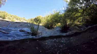 Etrafında bitki örtüsü olan su akıntısı. Kayalar var. Çimler yeşil.