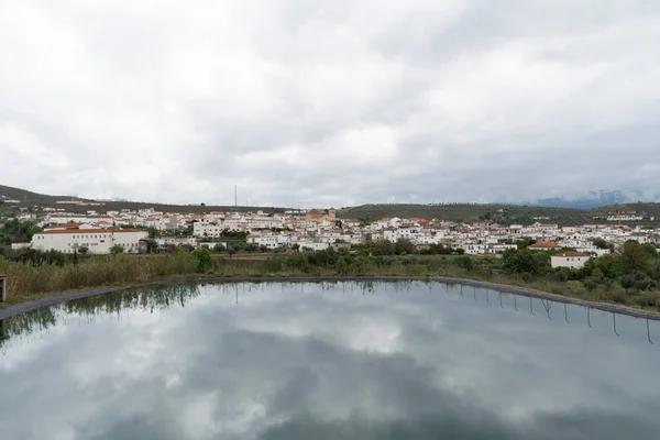 water reservoir next to a city, in the water the cloudy sky is reflected, the town has a church and the houses are painted white