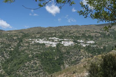 Dağın kenarındaki köy yeşil ağaçlarla çevrili, gökyüzünde bulutlar var.