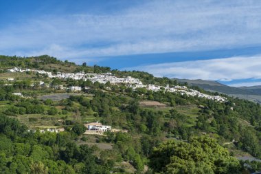 Dağın kenarındaki köy yeşil ağaçlar ve tarım arazileriyle çevrili, gökyüzü bulutlu.