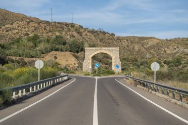 Yolda bir kavşakta dekoratif su kemeri var, yanında çalılar var ve yer çimlerle kaplı, arkasında ağaçlar ve dağlar var, gökyüzünde bulutlar var.