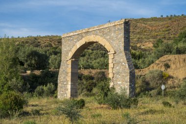 Yolda bir kavşakta dekoratif su kemeri var, yanında çalılar var ve yer çimlerle kaplı, arkasında ağaçlar ve dağlar var, gökyüzünde bulutlar var.