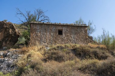 Ağaçlar ve bitki örtüsü arasında eski bir çiftlik evi, toprak örtüsü kuru, arkasında dağlar var, çiftlik evinin yanında büyük bir kaya var ve gökyüzü mavi
