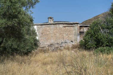 Ağaçlar ve bitkiler arasındaki eski çiftlik evi, toprak bitkisi kuru, arkasında dağlar var ve gökyüzü mavi.
