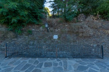 Ağaçlı taş duvar, metal bir korkuluk var, duvarda bitki örtüsü ve lamba direği var, tabakta Pileta çeşmesi yazıyor.