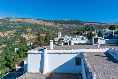 Güney İspanya 'nın dağlık bölgesi, dağ bitki ve ağaçlarla kaplı, bir kasabanın evleri görülüyor ve gökyüzünde bulutlar var.