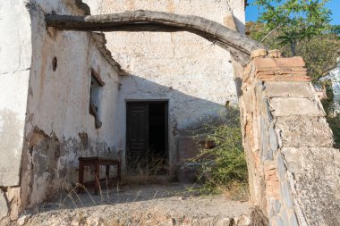Terk edilmiş çiftlik evi, ön cephesi beyaz ve pencereleri ve kapıları var, oturulacak ahşap bir bank var, koridordan geçen 2 çubuk.