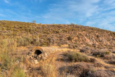 Dağ, bitki örtüsüyle, gökyüzü bulutlarla kaplı. Girişinde taşlar olan küçük bir tünel var. Dağın tepesinde, manzaralı bir korkuluk.