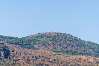 Ağaçlarla kaplı dağlık bir arazi, dağın tepesinde bir muhafız kulübesi ve metal bir direk var, gökyüzü temiz.