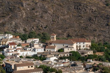 Beyaz cepheli ve kiliseli küçük bir kasaba. Kasabanın etrafında zeytin ağaçları ve arkasında bir dağ var.