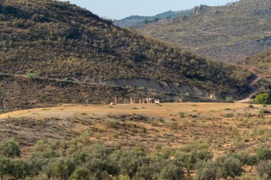 Dağlık alanda bitki örtüsü var, zeytin ağaçları yetiştiriliyor, harabeler içinde bir çiftlik evi var, gökyüzü açık.