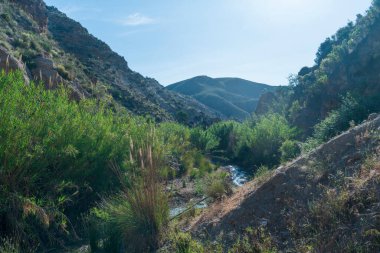 Nehir dağlık bir alanı geçiyor, nehir su taşıyor ve bitki örtüsü var, gökyüzü açık.