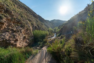 Nehir dağlık bir alanı geçiyor, nehir su taşıyor ve bitki örtüsü var, gökyüzü açık.