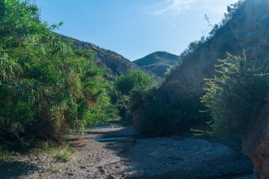 Nehir dağlık bir alanı geçiyor, nehir su taşıyor ve bitki örtüsü var, gökyüzü açık.