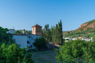 Küçük bir kasabadaki bir kilisenin kulesi, kule tuğlalardan yapılmıştır, evler beyaza boyanmıştır, etrafında dağlar vardır ve gökyüzü temizdir.