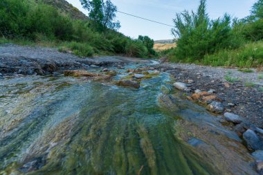 Nehirden akan su, küçük çalılar ve sazlıklar, gökyüzünde bulutlar, yerde kum ve taşlar var.